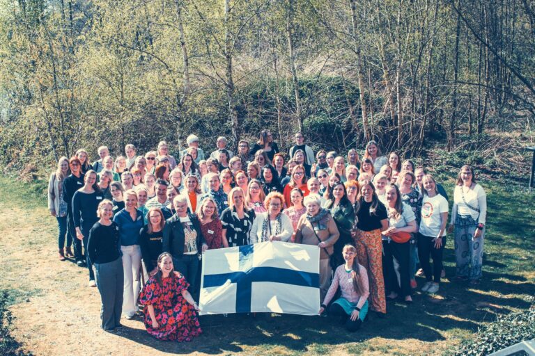 Joukko suomalaisia naisia ulkona. On kevät. Edessä istuvat pitävät kiinni isosta Suomen lipusta.