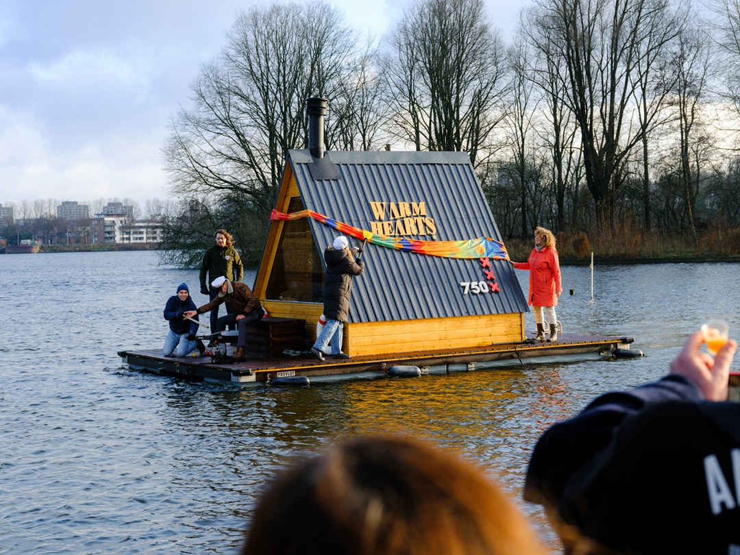 Warm Hearts -saunalautta kelluu Sloterplas-järvellä Amsterdamissa. Lautalla seisoo ja istuu useita ihmisiä värikkäissä talvivaatteissa. Taustalla on paljaita puita ja kaukaisia rakennuksia veden äärellä.