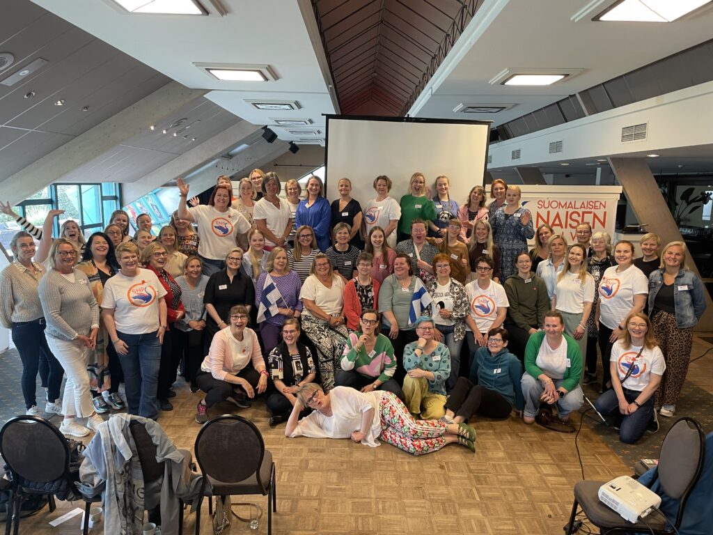 Joukko suomalaisia naisia kokoontunut ryhmäkuvaan sisätilassa. Osalla kädessään Suomen lippuja. Taustalla juliste Suomalaisen Naisen päivät.