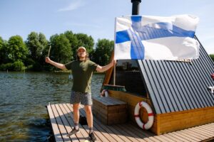 Hymyilevä mies seisoo Warm Hearts Saunan saunalautalla, kädessään Suomen lippu ja saunavihta. Taustalla vettä, vehreitä puita ja sininen taivas.