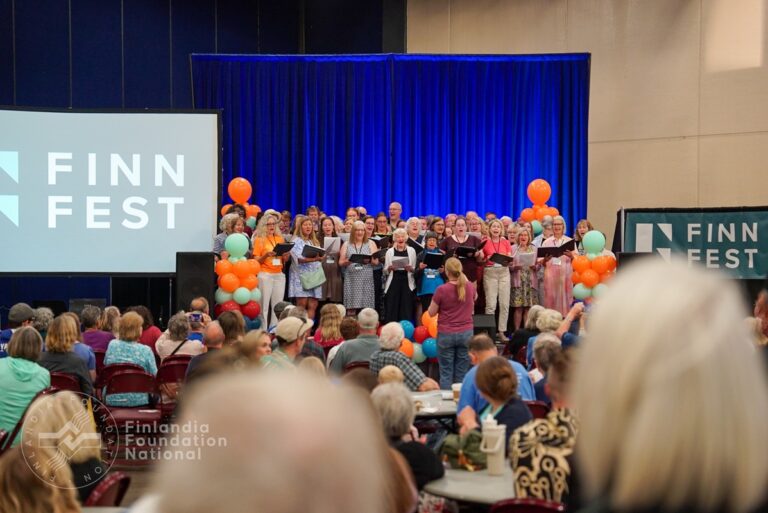 Ulkosuomalaiskuoro esiintyy lavalla ja sali on täynnä yleisöä. Lavan taustalla on suuri näyttö, jossa lukee Finn Fest.