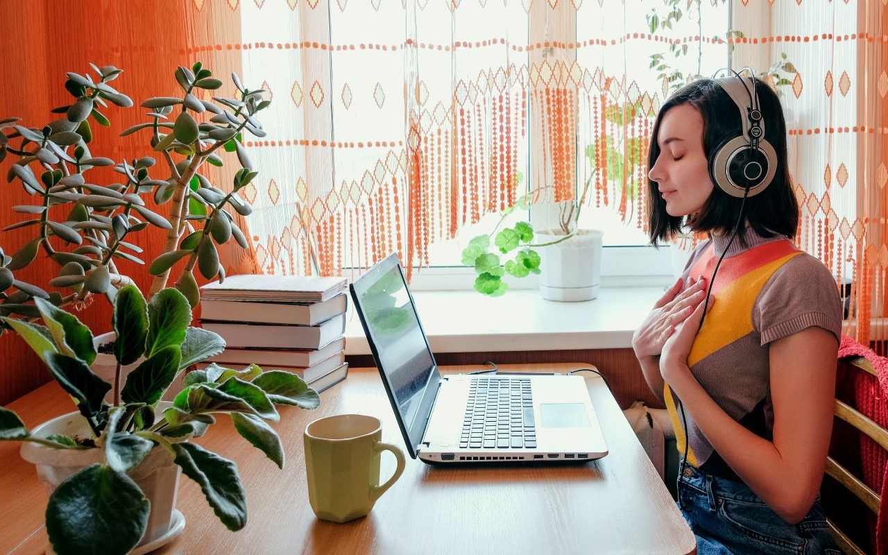 Nainen istuu pöydän ääressä tietokone edessään, kuulokkeet korvillaan ja kädet rintakehällään silmät kiinni.