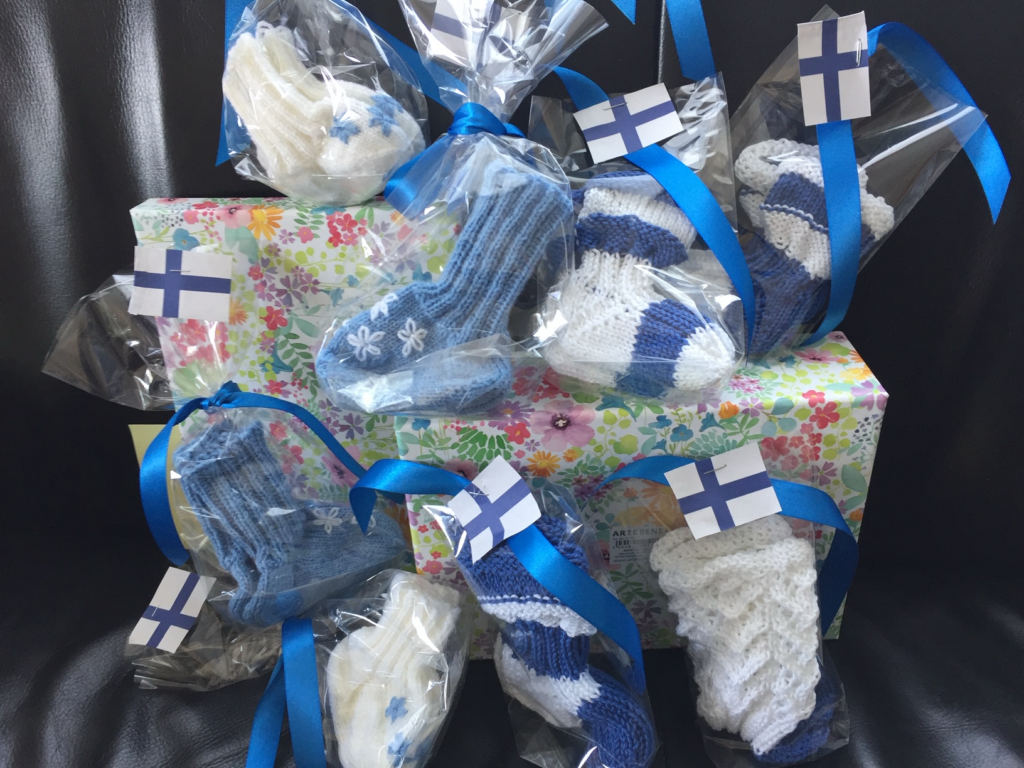 Sinivalkoisin nauhoin koristeltuja käsinneulottuja lasten villasukkia ja lapasia sellofaanipakkauksissa, aseteltuna lahjapaketin päälle.